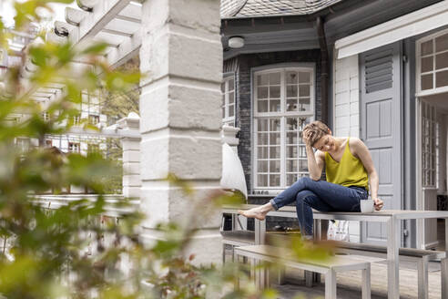 Relaxed woman sitting on terrace table in garden of her home - MJFKF00005