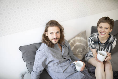 Porträt eines glücklichen jungen Paares im Pyjama auf der Couch zu Hause - MJFKF00015