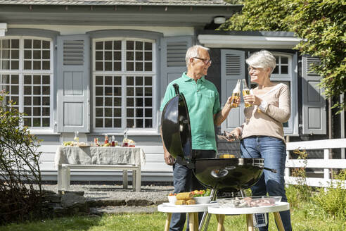 Glückliches älteres Paar beim Grillen im Garten ihres Hauses, das mit Bierflaschen anstößt - MJFKF00046