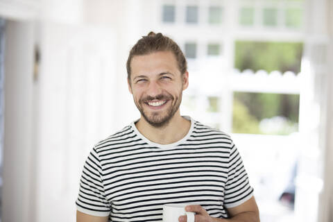 Porträt eines lächelnden jungen Mannes, der eine Tasse Kaffee zu Hause hält, lizenzfreies Stockfoto