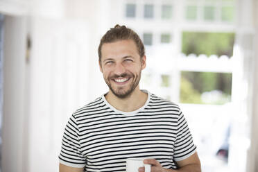 Portrait of smiling young man holding cup of coffee at home - MJFKF00061