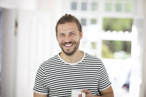 Porträt eines lächelnden jungen Mannes, der eine Tasse Kaffee zu Hause hält - MJFKF00061