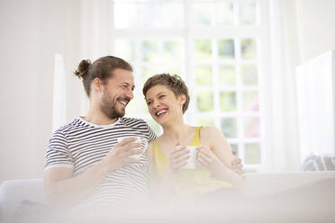 Glückliches junges Paar mit Tassen auf der Couch zu Hause - MJFKF00062