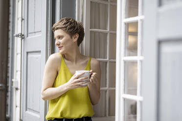 Smiling woman with cup of coffee at the terrace door - MJFKF00066