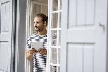 Young man with tablet standing at terrace door at home - MJFKF00068