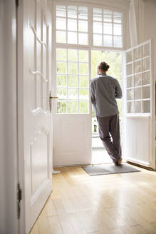 Young man in pyjama standing at the terrace door at home - MJFKF00088