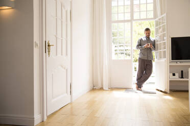 Young man in pyjama standing at the terrace door at home - MJFKF00090