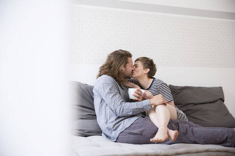 Junges Paar im Pyjama küsst sich auf der Couch zu Hause, lizenzfreies Stockfoto