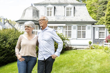 Smiling senior couple standing in garden of their home - MJFKF00123