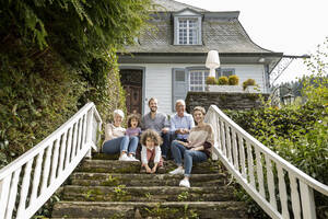 Eine glückliche Großfamilie sitzt auf einer Treppe im Garten ihres Hauses - MJFKF00136