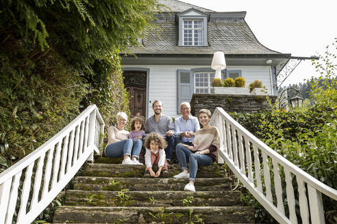 Happy extended family sitting on stairs in garden of their home - MJFKF00136