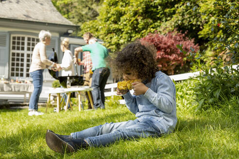 Junge isst einen Maiskolben auf einem Familiengrill im Garten - MJFKF00145
