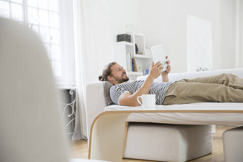 Junger Mann liegt zu Hause auf der Couch und benutzt ein Tablet, lizenzfreies Stockfoto