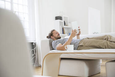 Junger Mann liegt zu Hause auf der Couch und benutzt ein Tablet - MJFKF00176