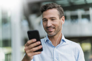 Smiling businessman with earphones and cell phone in the city - DIGF08435