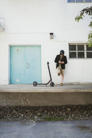 Mann mit Elektroroller lehnt an Fassade und benutzt Smartphone, lizenzfreies Stockfoto