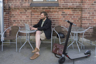 Mann mit Elektroroller sitzt in einem Straßencafé und benutzt einen Laptop - PSIF00320
