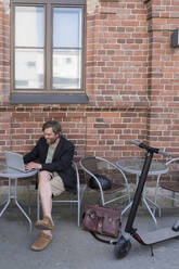 Mann mit Elektroroller sitzt in einem Straßencafé und benutzt einen Laptop - PSIF00321