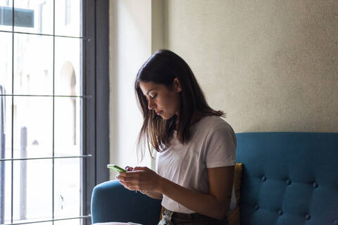 Young woman sitting on a couch at the window using cell phone stock photo