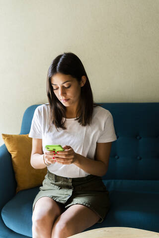 Junge Frau sitzt auf einer Couch und benutzt ein Mobiltelefon, lizenzfreies Stockfoto