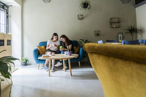 Two young female friends sitting on a couch in a cafe sharing cell phone stock photo