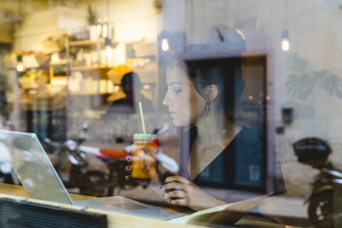 Junge Frau mit Smoothie und Laptop hinter einer Fensterscheibe in einem Cafe, lizenzfreies Stockfoto