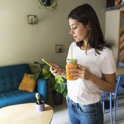 Junge Frau mit einem Smoothie und einem Mobiltelefon in einem Café - GIOF07098