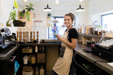 Portrait of smiling young woman with cell phone behind the counter in a cafe - GIOF07101