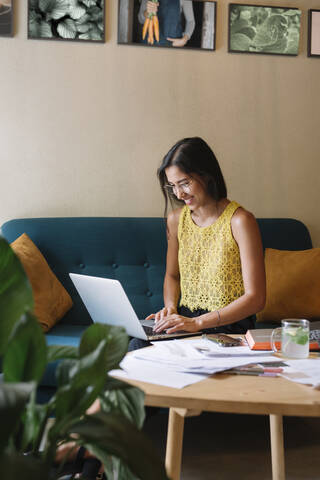 Lächelnde Modedesignerin sitzt auf der Couch und benutzt einen Laptop, lizenzfreies Stockfoto