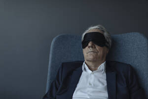 Älterer Geschäftsmann sitzt im Sessel und trägt eine Augenmaske - GUSF02534