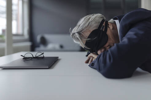 Senior businessman wearing eye mask lying on desk - GUSF02546