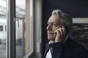 Porträt eines älteren Geschäftsmannes, der am Fenster mit einem Handy telefoniert - GUSF02576
