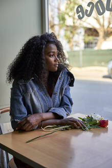 Portrait of young African woman with flowers on the table in a cafe, looking out of window - VEGF00688