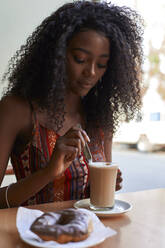 Porträt einer jungen afrikanischen Frau, die in einem Café einen Kaffee und einen Doughnut trinkt - VEGF00695