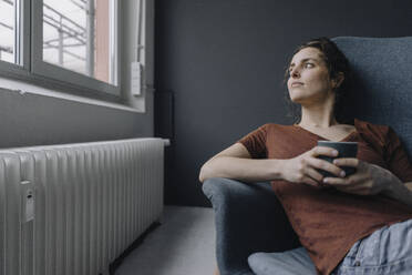 Junge Frau mit einer Tasse Kaffee auf einem Liegestuhl und Blick aus dem Fenster - KNSF06558