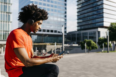 Junger Sportler sitzt auf einer Bank, benutzt sein Smartphone und hört Musik - JRFF03722