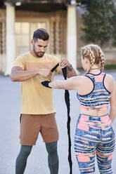 Fitnesstrainer, der mit einer jungen Frau im Freien in der Stadt Boxunterricht gibt und einen Verband anlegt - JNDF00093