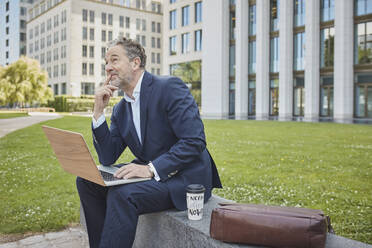 Mature businessman sitting on a wall in the city using laptop - RORF01852