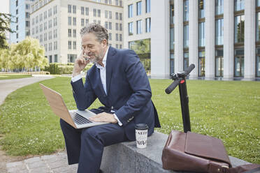 Mature businessman sitting on a wall in the city using laptop - RORF01853
