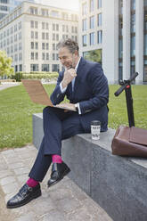 Happy mature businessman sitting on a wall in the city using laptop - RORF01854