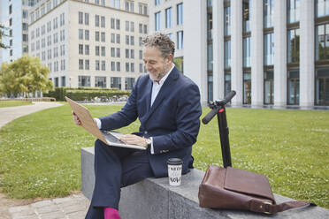 Glücklicher reifer Geschäftsmann sitzt auf einer Mauer in der Stadt und benutzt einen Laptop - RORF01855