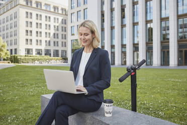 Geschäftsfrau sitzt auf einer Mauer in der Stadt und benutzt einen Laptop - RORF01874