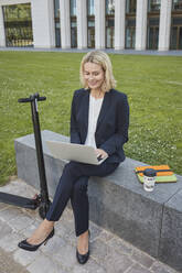 Businesswoman sitting on a wall in the city using laptop - RORF01875