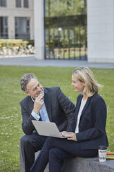 Businesswoman and businessman sitting on a wall in the city using laptop - RORF01877