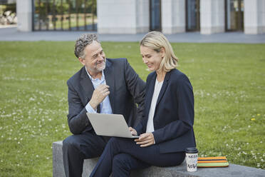 Geschäftsfrau und Geschäftsmann sitzen auf einer Mauer in der Stadt und benutzen einen Laptop - RORF01878