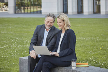 Businesswoman and businessman sitting on a wall in the city using laptop - RORF01879