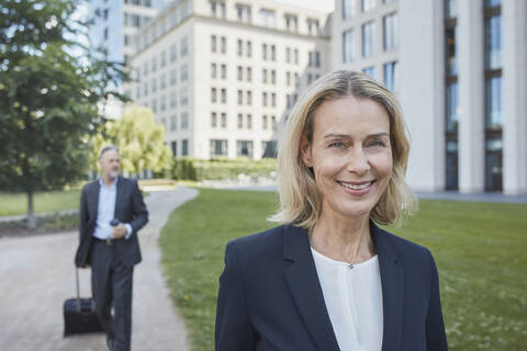 Porrait einer lächelnden blonden Geschäftsfrau in der Stadt mit einem Geschäftsmann im Hintergrund, lizenzfreies Stockfoto