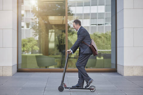 Geschäftsmann auf E-Scooter beim Passieren eines Bürogebäudes in der Stadt, lizenzfreies Stockfoto