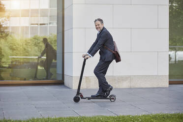 Businessman on e-scooter passing office building in the city - RORF01887