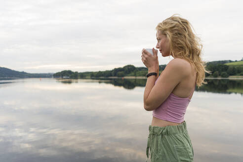 Junge Frau trinkt Kaffee aus einer Tasse an einem See - JOSF03602